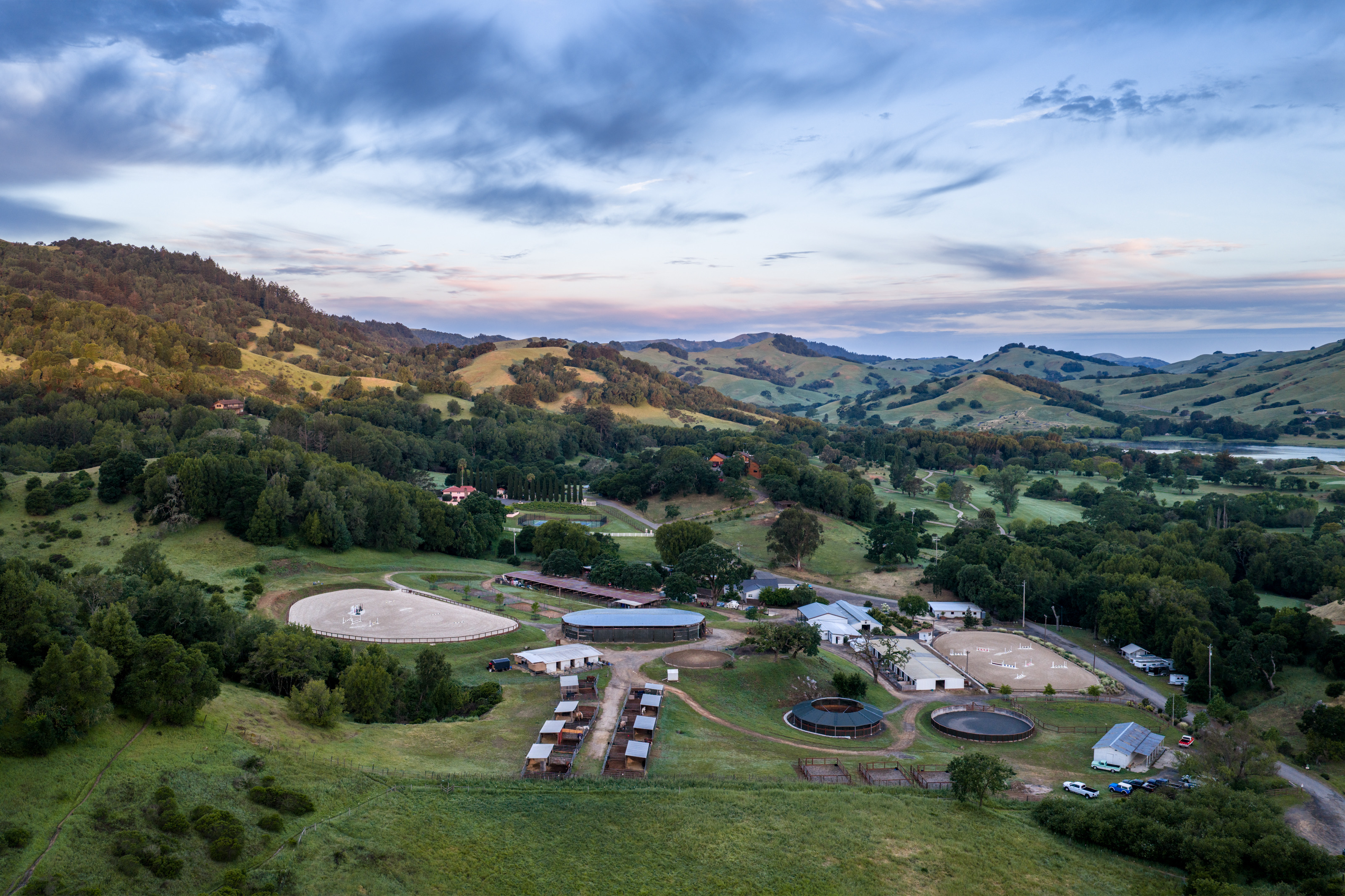 The farm’s barns and arenas below rolling hills, with a lake in the distance at dusk.