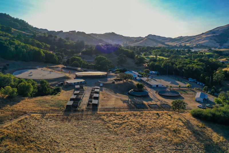Backlit golden-hour aerial showing the arenas, round pens, stalls, and wooded hills