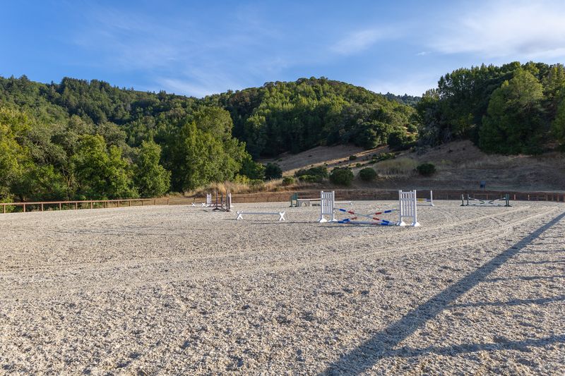 Outdoor sand jump arena with a crossrail and wooded hills beneath a clear blue sky
