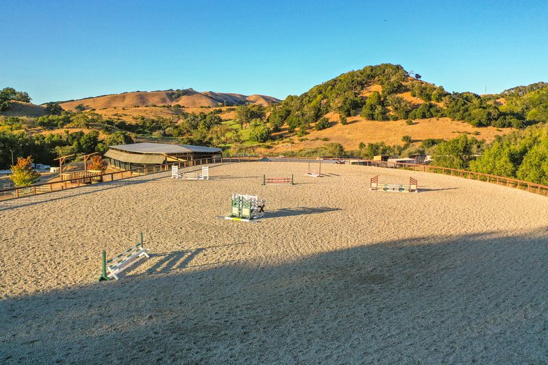 Outdoor jump arena at golden hour with the covered arena and wooded hillside beyond
