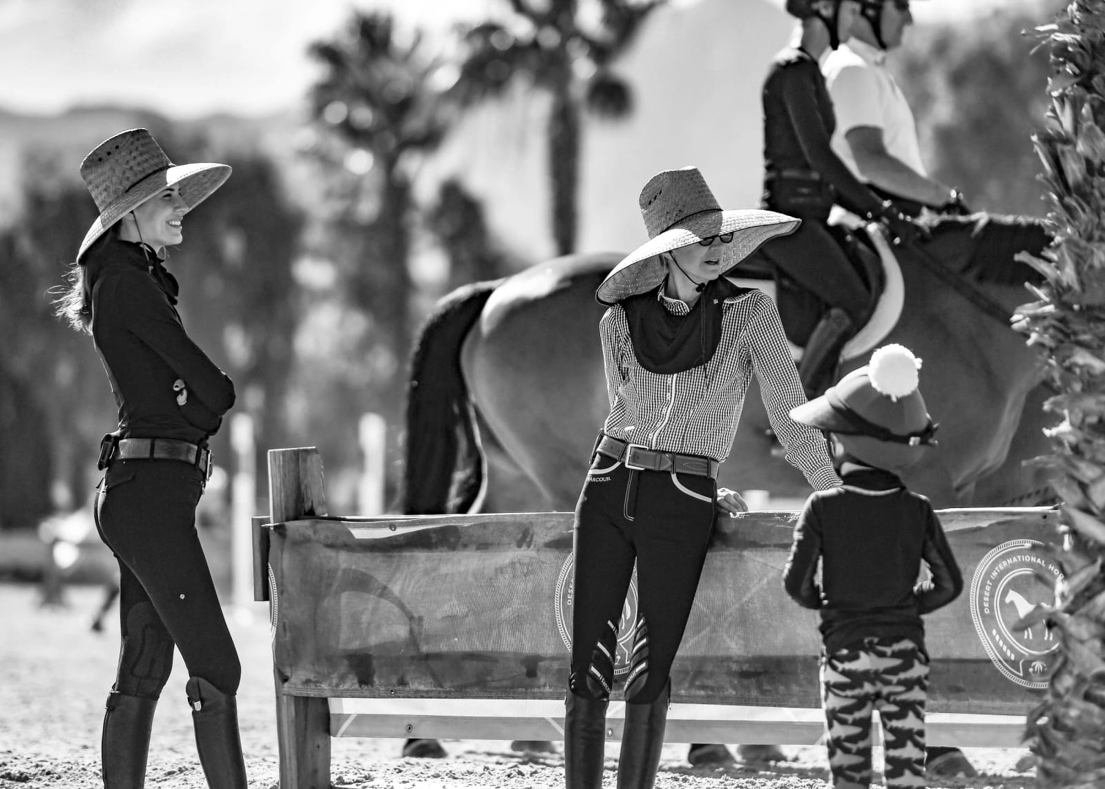 Two women in wide straw hats and a young child stand at the rail of a show ring, with a horse and rider behind them.